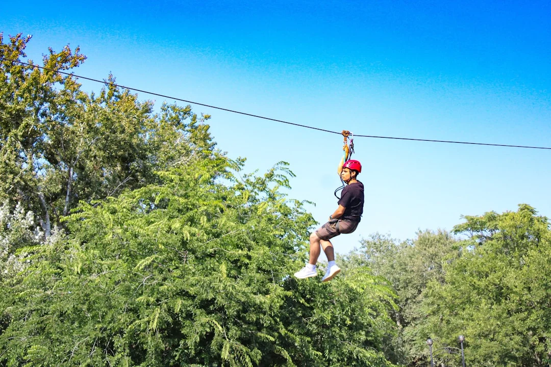 A smiling child in a helmet and harness flying along a backyard zipline strung between two mature oak trees on a sunny afternoon, with a parent watching from below